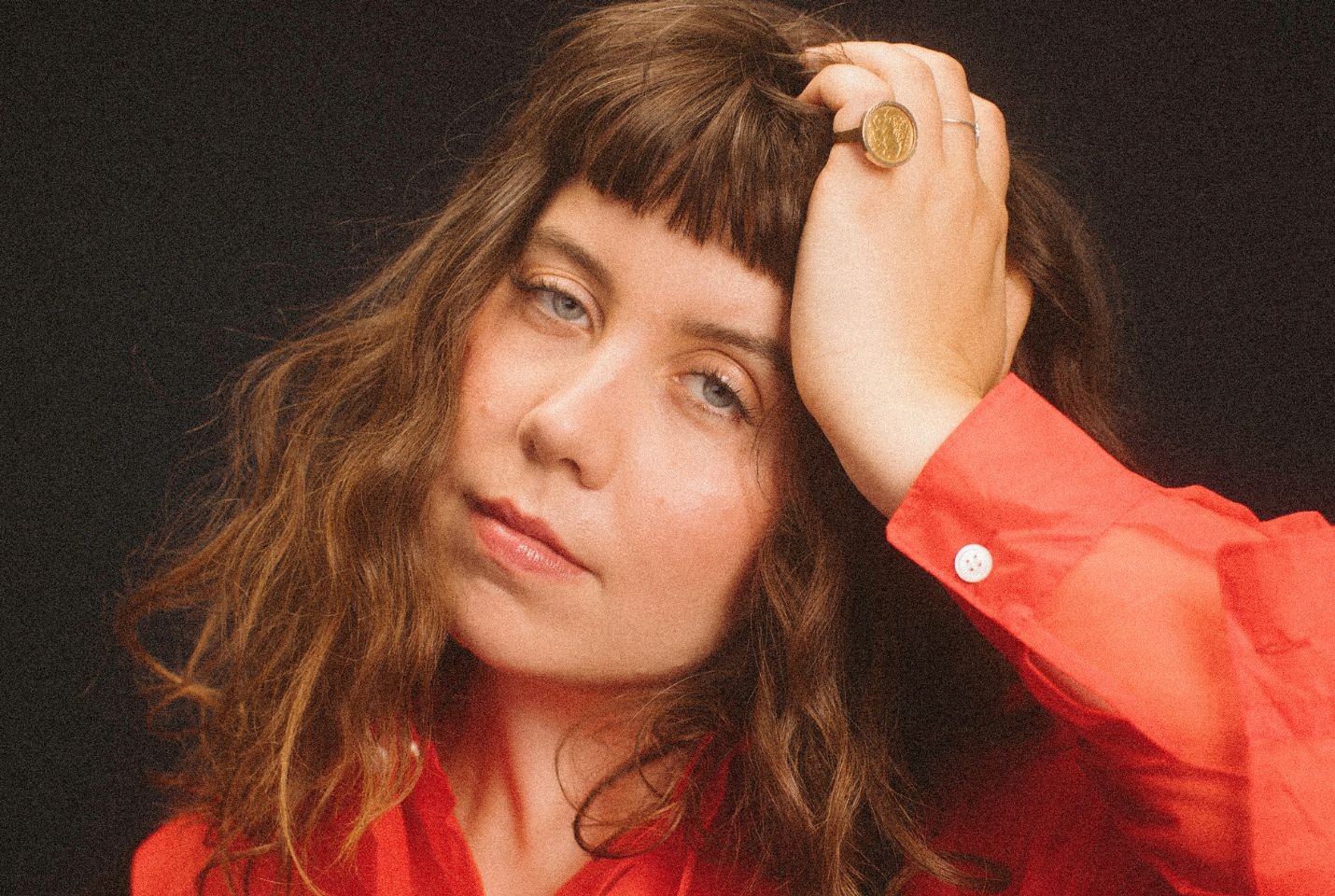A woman wearing a red shirt with her hand rested on her head. She has a large gold coin ring on her pinky finger. She has a curly brown bob with a fringe